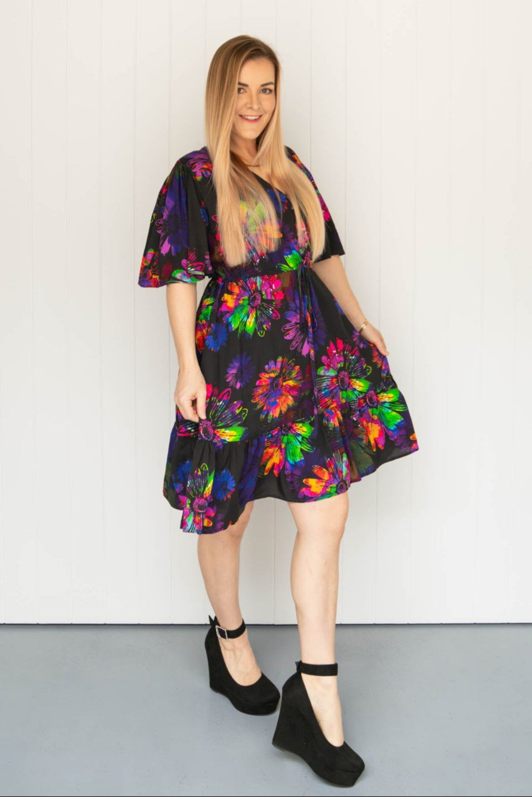 A model wearing a multicolored mini dress with a floral pattern, featuring a V-neckline, wide sleeves, a drawstring waistband, and a tiered hem.