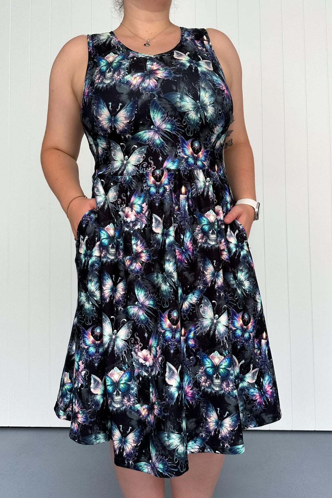 A sleeveless knee-length dress with a printed design featuring butterflies and gothic motifs, held by a model with hands in the deep side pockets.