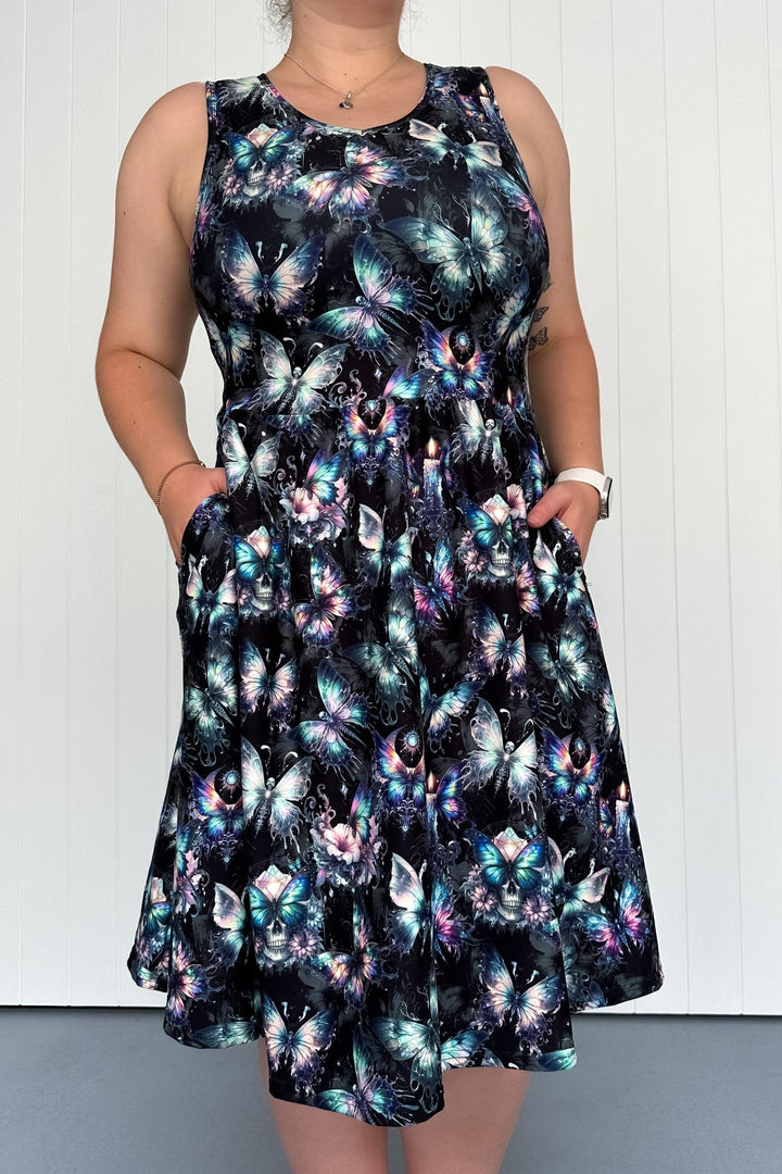 A sleeveless knee-length dress with a printed design featuring butterflies and gothic motifs, held by a model with hands in the deep side pockets.