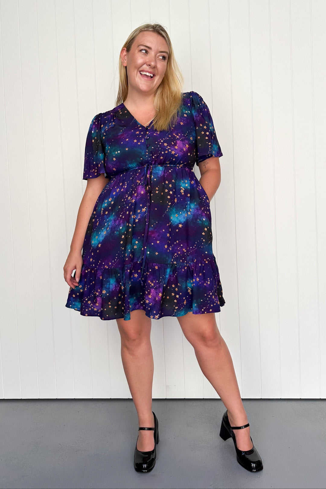 A model wearing a mini dress with a relaxed fit, tiered hem, and a universe shimmer print consisting of purples and blues with gold stars.