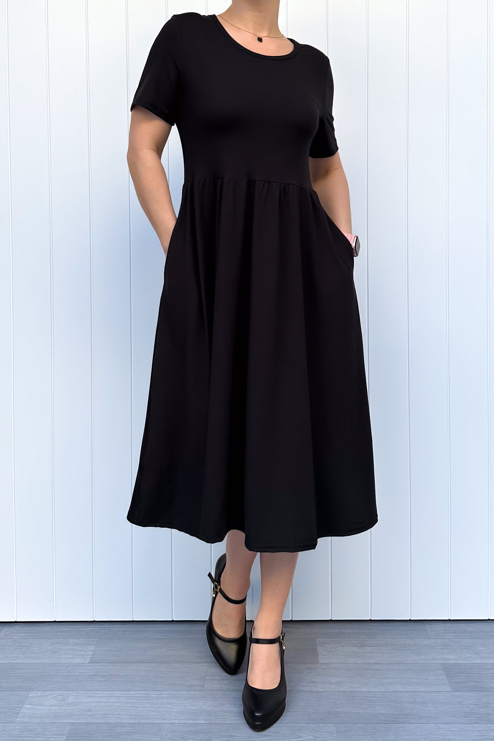Woman wearing a black, short sleeve, midi length dress standing against a white wooden panel background