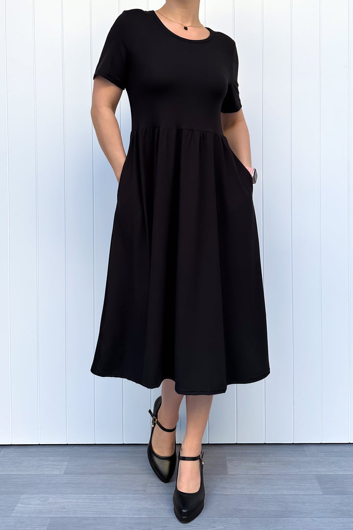 Woman wearing a black, short sleeve, midi length dress standing against a white wooden panel background