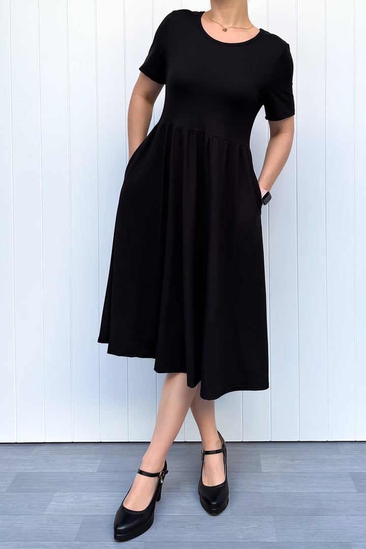 Woman wearing a black, short sleeve, midi length dress standing against a white wooden panel background
