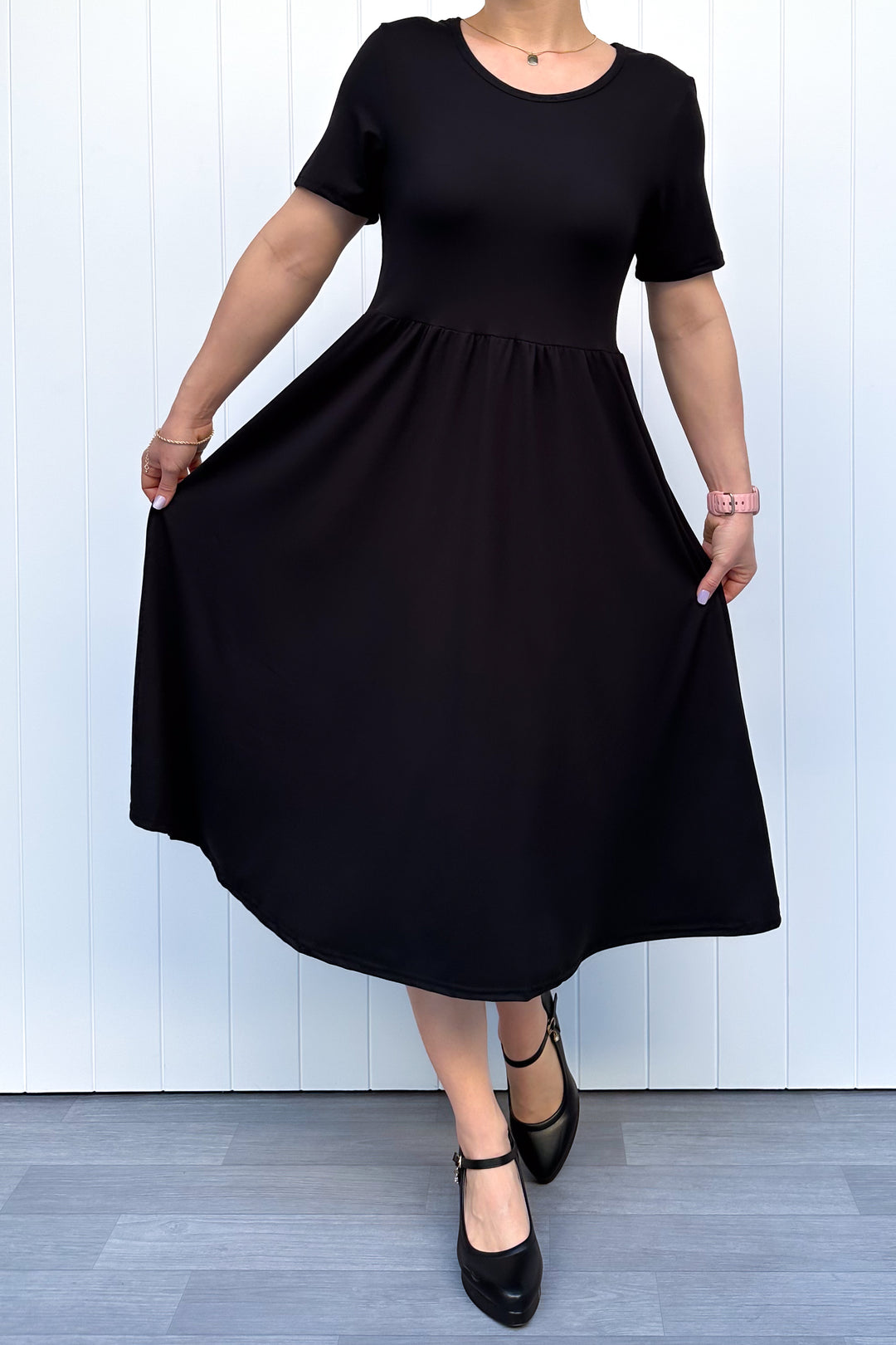 Woman wearing a black, short sleeve, midi length dress standing against a white wooden panel background