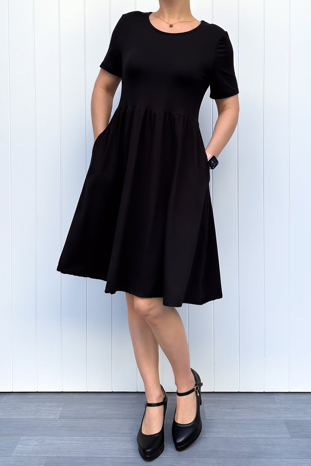 Woman wearing a black, short sleeve, knee length dress standing against a white wall