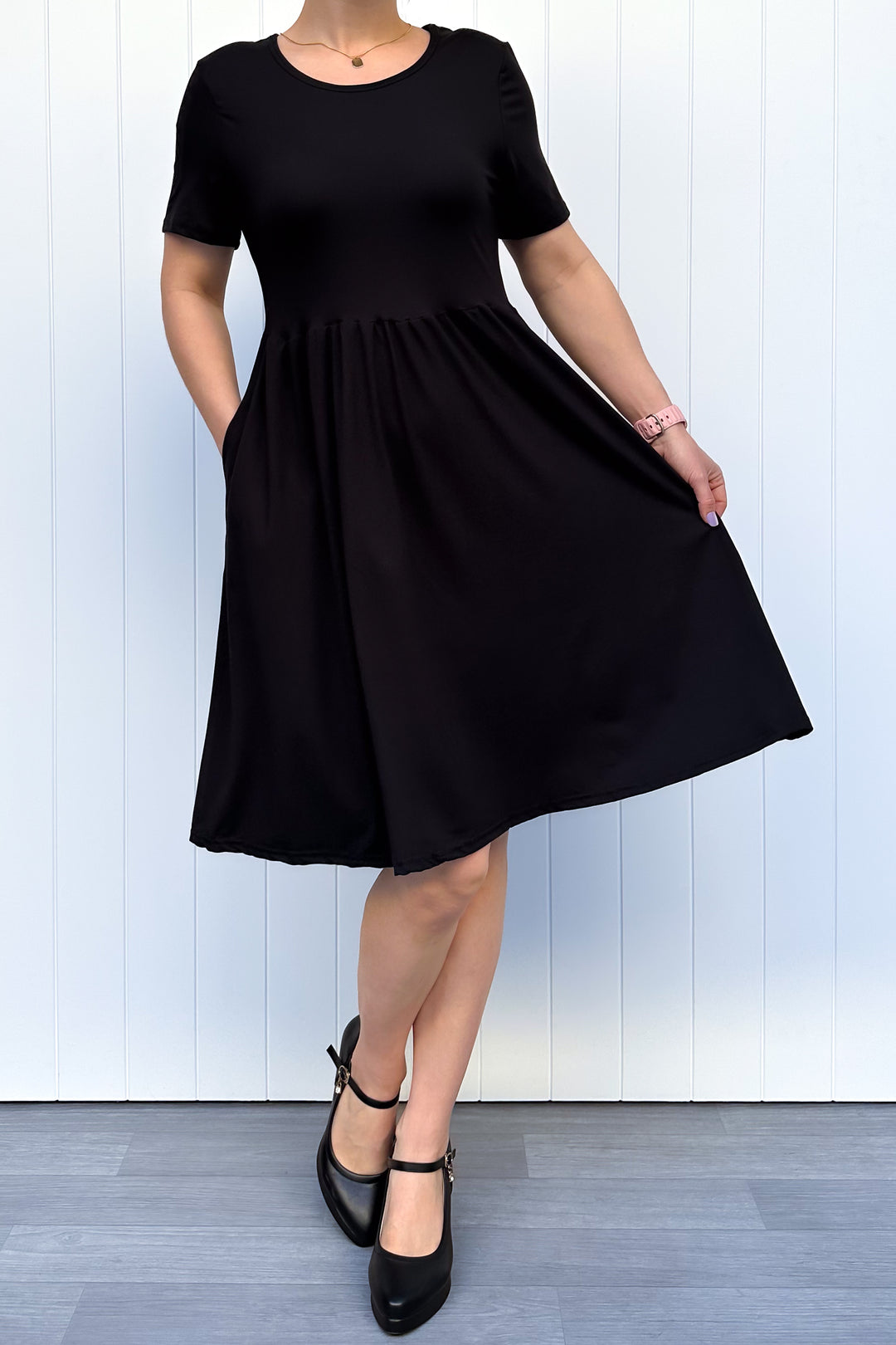 Woman wearing a black, short sleeve, knee length dress standing against a white wall