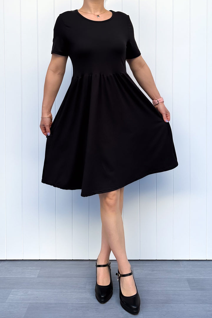 Woman wearing a black, short sleeve, knee length dress against a white wooden panel background