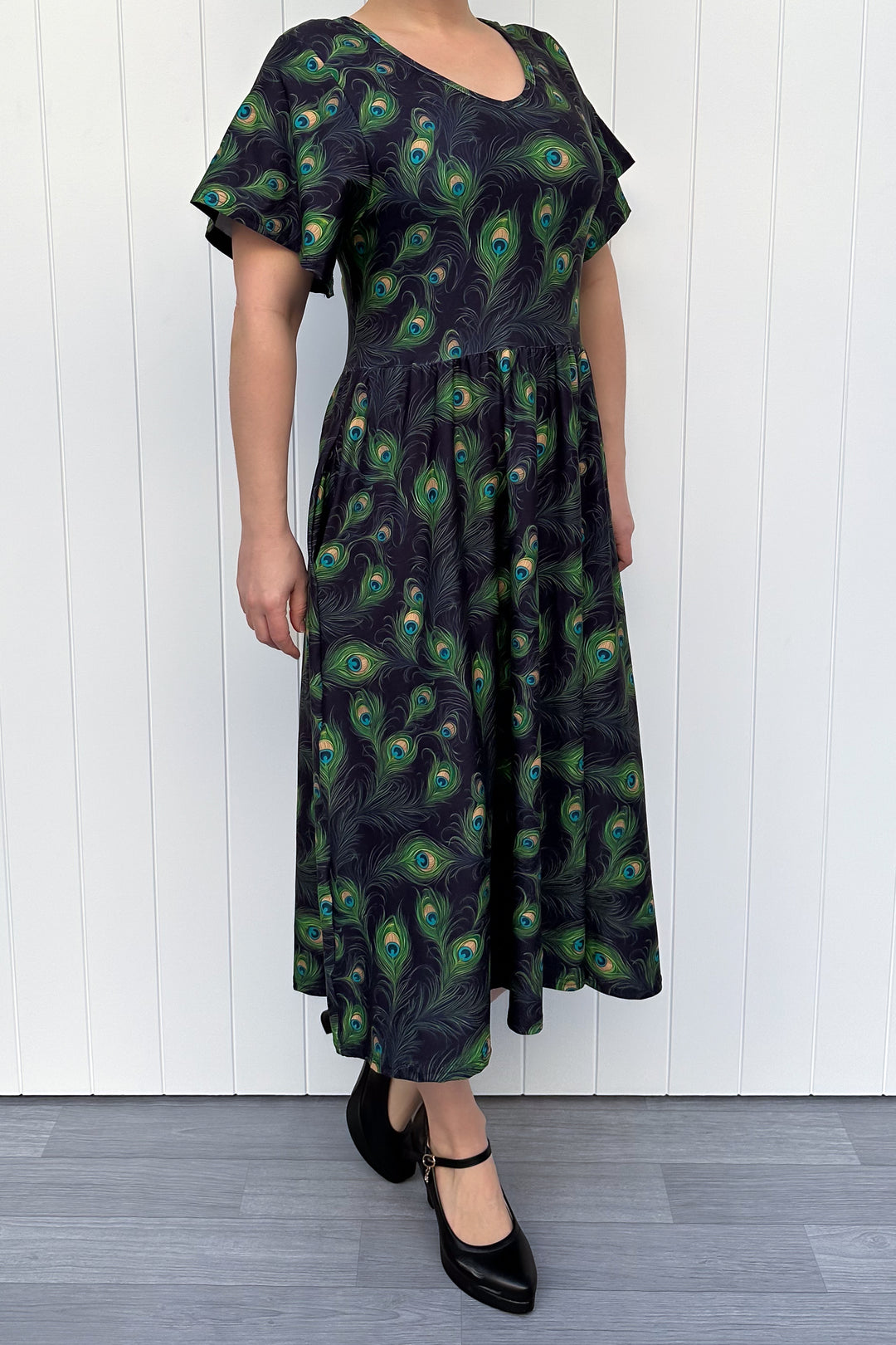 Person wearing a black dress with peacock feather pattern against a white wooden panel background
