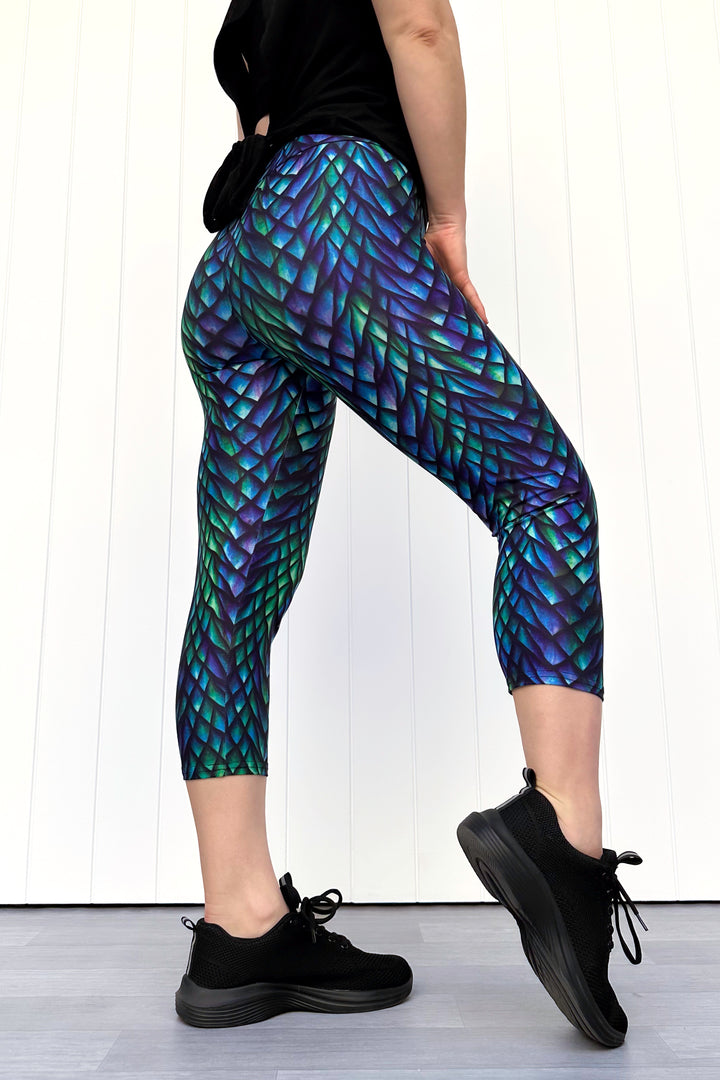 Person wearing green and purple capri leggings with a dragon scale pattern, standing against a white wall.